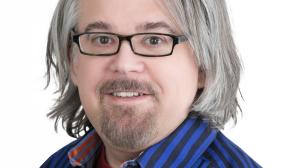 A man with long gray hand and black glasses, wearing a blue striped shirt. He has a goatee.