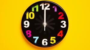 A black wall clock on a bright yellow background with colorful numbers and spinning hands.