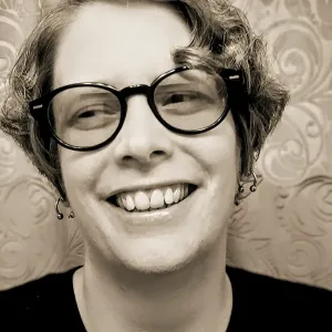 Black and white head shot photo of author Mattie-Bretton Hughes. He is smiling with is head tilted slightly to the left. He has short wavy blonde hair and is wearing glasses. The background is a soft grey curtain. 