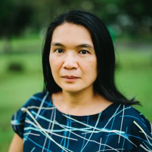 A headshot of an Asian woman with shoulder-length straight black hair in a blue top with crisscrossing stripes, taken outdoors in a park