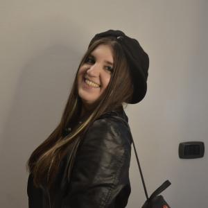 Maja is a white girl with long brown hair. In this picture she is smiling towards the camera, her body is turned on the side. She's standing against a white wall and is wearing a leather jacket and a blue greek hat.