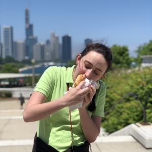 A woman in a light green polo using both hands to take a large bite of a wrapped hot dog.